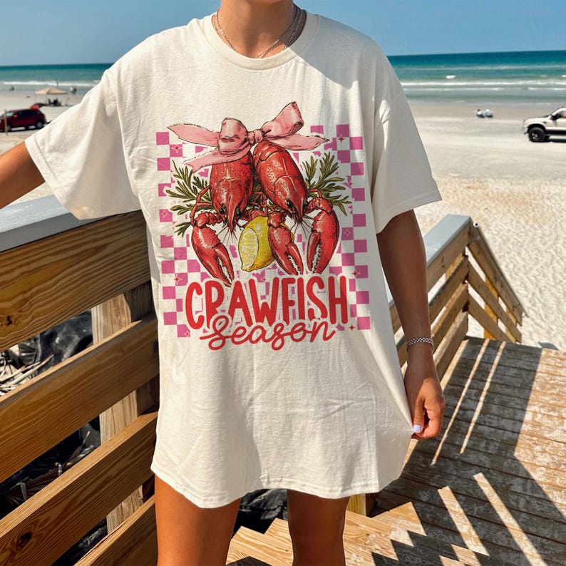 May include: Off-white t-shirt featuring a graphic of red crawfish, a lemon slice, and a pink bow on a pink and white checkered background. The text "CRAWFISH Season" is below the graphic. The shirt is a casual, oversized style.
