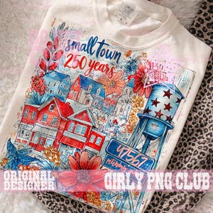 May include: Off-white t-shirt with a vibrant print of a small town scene, including red and blue houses, a water tower, and floral details. The text "small town 250 years" is above the town illustration. A street sign displays the number "47567".