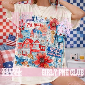 May include: A cream-coloured t-shirt with a vibrant illustration of a small town, featuring red, blue, and white buildings, flowers, and a water tower. The text "small town 250 years" is displayed. The town's postcode, 47567, is also visible.