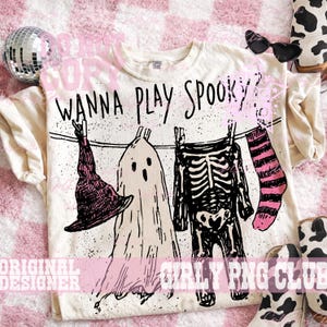 Op de afbeelding: Een cr&egrave;mekleurig t-shirt met een zwarte afbeelding van een waslijn met een heksenhoed, een geest, een skeletromp en een gestreepte sok. De tekst "WANNA PLAY SPOOKY?" staat in een zwart handschrift boven de waslijn gedrukt.