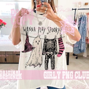 Op de afbeelding: Een wit t-shirt met korte mouwen en een zwarte afbeelding van een waslijn met een heksenhoed, een spook, een skelettenpak en een gestreepte sok. De tekst "WANNA PLAY SPOOKY?" is in het zwart boven de illustratie gedrukt.