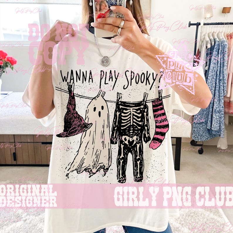 Op de afbeelding: Een wit t-shirt met korte mouwen en een zwarte afbeelding van een waslijn met een heksenhoed, een spook, een skelettenpak en een gestreepte sok. De tekst "WANNA PLAY SPOOKY?" is in het zwart boven de illustratie gedrukt.