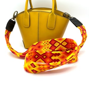 May include: A small, yellow leather handbag with a top handle and a long, colourful woven strap. The strap is red, orange, and yellow with a geometric pattern.