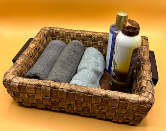 May include: A woven wicker basket with brown wooden handles, containing two gray towels, a blue towel, a clear bottle with a blue label, and a white bottle with a gold lid.