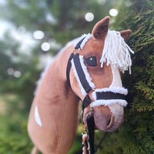 Può includere: Una testa di cavallo di peluche marrone e bianca con un morsetto nero e una corda di piombo. Il cavallo ha una criniera e una coda bianche.