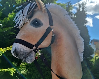 May include: A tan hobby horse with a black bridle and white and black mane. The toy horse has a wooden stick for a body and is set against a backdrop of green foliage and a blue sky.