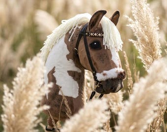 May include: A hobby horse with a brown and white spotted coat, featuring a cream-colored yarn mane and a black bridle with metallic stud accents. The horse head is mounted on a wooden stick, showcasing dark eyes and a soft, plush texture.