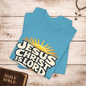 May include: A light blue t-shirt featuring the text "JESUS CHRIST IS LORD" in a retro font, with a yellow sunburst design. A brown leather-bound Holy Bible and a cross necklace are also visible.