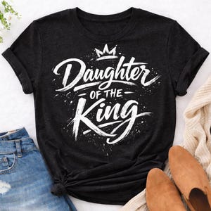 May include: Dark gray t-shirt with the text "Daughter of the King" in white script. The design includes a crown and splatter effects. The shirt is folded, with blue jeans and brown shoes visible.