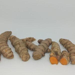 May include: Close-up of several turmeric root pieces, some with the skin on and others cut open to reveal the bright orange flesh. The roots are arranged on a white background.