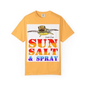 Pode incluir: Camiseta amarelo mostarda com um gráfico de uma tartaruga marinha acima das palavras "SUN SALT & SPRAY" em um gradiente de laranja, amarelo e azul. A camiseta tem gola redonda e mangas curtas.