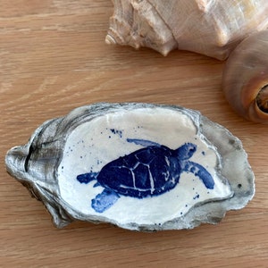May include: A blue and white sea turtle design painted on a gray oyster shell. The shell is a natural, organic shape and is perfect for holding small items.