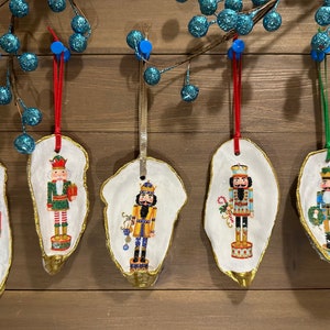 May include: Five handmade Christmas ornaments featuring nutcracker designs painted on oyster shells. Each ornament is adorned with gold trim and a ribbon for hanging.