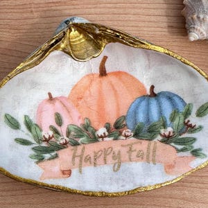 May include: A white seashell dish with a gold rim, painted with three pumpkins and green leaves. The pumpkins are orange, pink, and blue. The dish has a banner that says "Happy Fall".