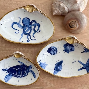 May include: Three white ceramic clam shells with gold trim, each featuring a different blue and white nautical design. One shell has an octopus, one has a sea turtle, and one has a starfish and seashells.