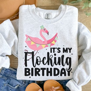 Puede incluir: Una sudadera gris con un flamenco rosa que lleva una corona de flores y sostiene una pancarta que dice "It's My Flocking Birthday".