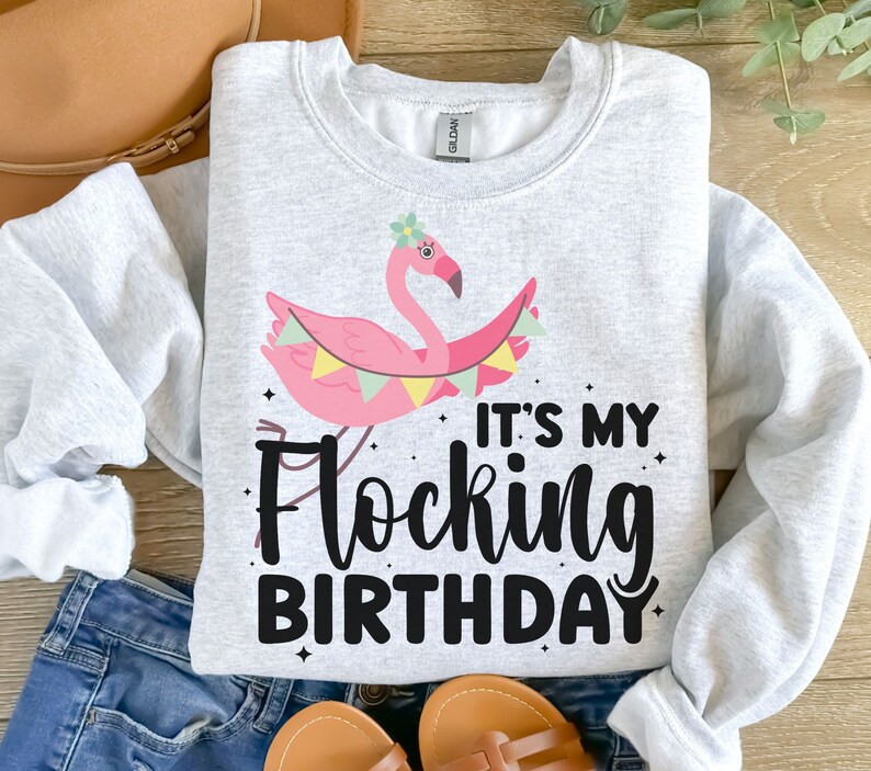 Puede incluir: Una sudadera gris con un flamenco rosa que lleva una corona de flores y sostiene una pancarta que dice "It's My Flocking Birthday".