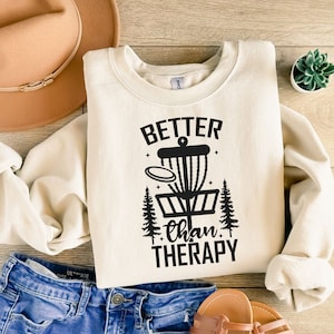 May include: A cream-colored sweatshirt with a black graphic of a disc golf basket and the text "Better than Therapy".
