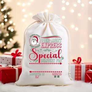 May include: White canvas gift bag with red and green text that reads "North Pole Post Office Over-Night Express Special Delivery For Inspected By Elf Checked". The bag has a drawstring closure and a small illustration of Santa Claus.