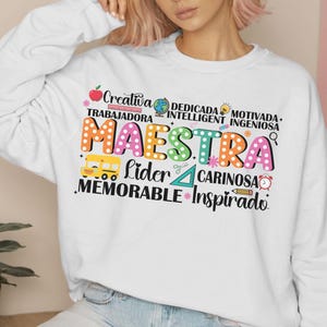 Pode incluir: Camisola branca com um design gr&aacute;fico colorido que diz "Maestra" e inclui palavras como "creativa", "trabajadora", "dedicada", "inteligente", "motivada", "ingeniosa", "lider", "cari&ntilde;osa", "memorable" e "inspirado".