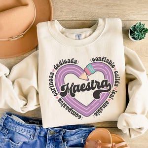 Pode incluir: Uma camisola de cor creme com um desenho de cora&ccedil;&atilde;o de l&aacute;pis roxo e a palavra "Maestra" numa fonte retro. O cora&ccedil;&atilde;o &eacute; rodeado por palavras em espanhol, incluindo "cari&ntilde;osa", "compastiva", "dedicada", "confiable", "amable", "leal" e "calida".