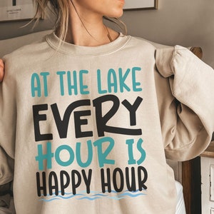 May include: A beige sweatshirt with the text "At the lake every hour is happy hour" in black and teal blue lettering. The text is surrounded by a wavy blue line.