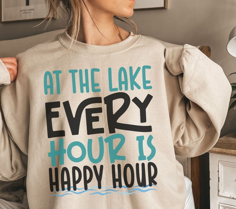May include: A beige sweatshirt with the text "At the lake every hour is happy hour" in black and teal blue lettering. The text is surrounded by a wavy blue line.