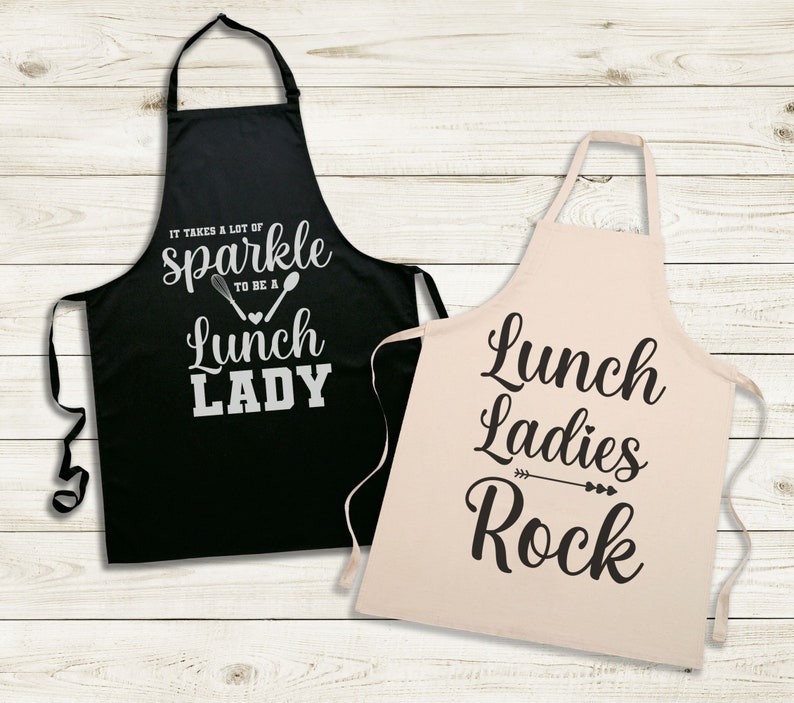 May include: Two black and white aprons with text. The black apron says "It takes a lot of sparkle to be a lunch lady". The white apron says "Lunch ladies rock".