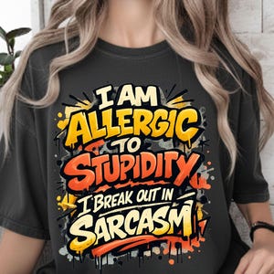 Pu&ograve; includere: T-shirt grigio scuro con la scritta "I AM ALLERGIC TO STUPIDITY I BREAK OUT IN SARCASM" in giallo, arancione e bianco. Il testo ha un design in stile graffiti con schizzi di vernice.
