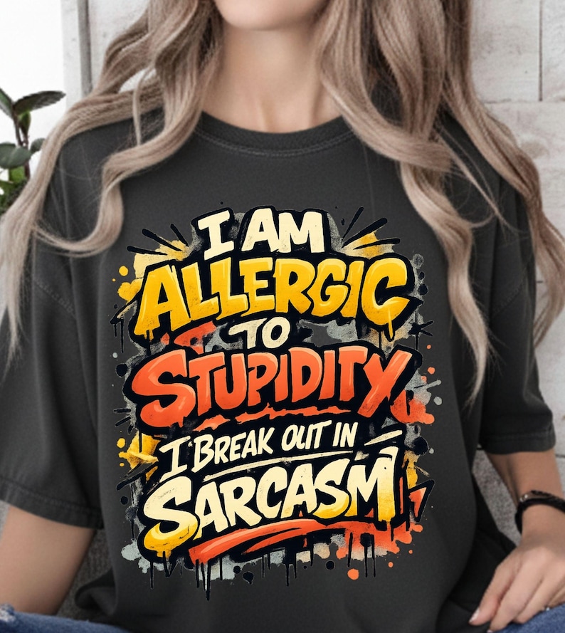 Pu&ograve; includere: T-shirt grigio scuro con la scritta "I AM ALLERGIC TO STUPIDITY I BREAK OUT IN SARCASM" in giallo, arancione e bianco. Il testo ha un design in stile graffiti con schizzi di vernice.