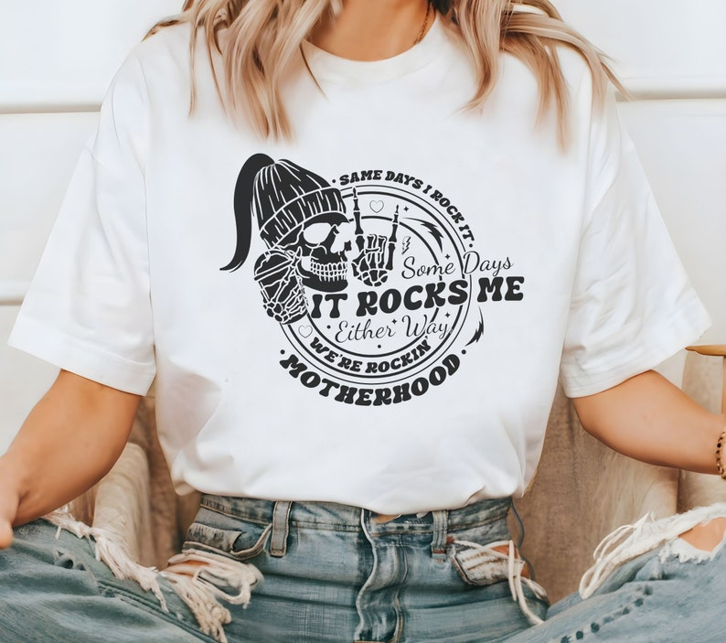 May include: White t-shirt with a black and white graphic design. The graphic features a skull wearing a beanie, holding a coffee cup, and making a rock and roll hand gesture. The text reads "Same days I rock it. Some days it rocks me. Either way we're rockin' motherhood."