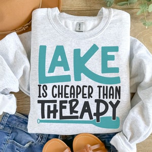 May include: A gray sweatshirt with the text "LAKE IS CHEAPER THAN THERAPY" in teal and black lettering. A teal paddle is depicted below the text.