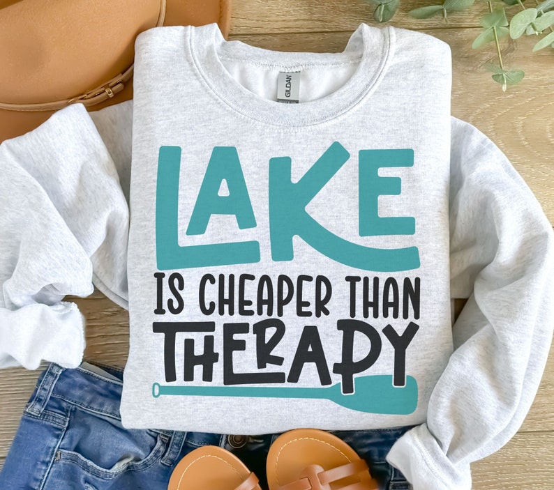 May include: A gray sweatshirt with the text "LAKE IS CHEAPER THAN THERAPY" in teal and black lettering. A teal paddle is depicted below the text.