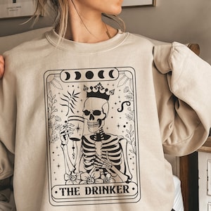May include: A beige sweatshirt with a black and white illustration of a skeleton holding a glass. The illustration is in the style of a tarot card and reads "The Drinker".
