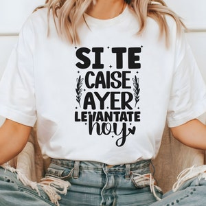 May include: White t-shirt with black text that reads "Si te caise ayer levantate hoy" with a small heart symbol. The text is surrounded by small black stars and a floral design.