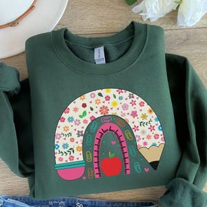 Pode incluir: Uma camisa de frio verde escura com um desenho de arco-&iacute;ris feito de um l&aacute;pis, uma r&eacute;gua, clipes e flores. O arco-&iacute;ris &eacute; rosa, azul e amarelo com uma ma&ccedil;&atilde; vermelha no centro.