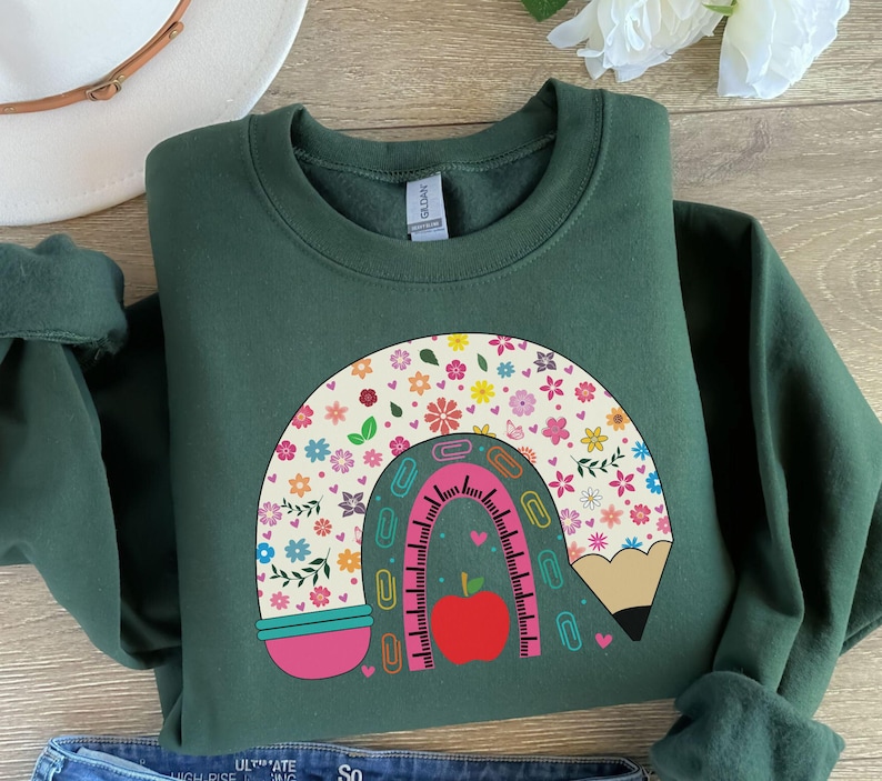 Pode incluir: Uma camisa de frio verde escura com um desenho de arco-&iacute;ris feito de um l&aacute;pis, uma r&eacute;gua, clipes e flores. O arco-&iacute;ris &eacute; rosa, azul e amarelo com uma ma&ccedil;&atilde; vermelha no centro.