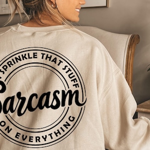 K&ouml;nnte beinhalten: Ein beiger Pullover mit einem schwarzen Kreis-Motiv, auf dem "I sprinkle that stuff Sarcasm on everything" steht.