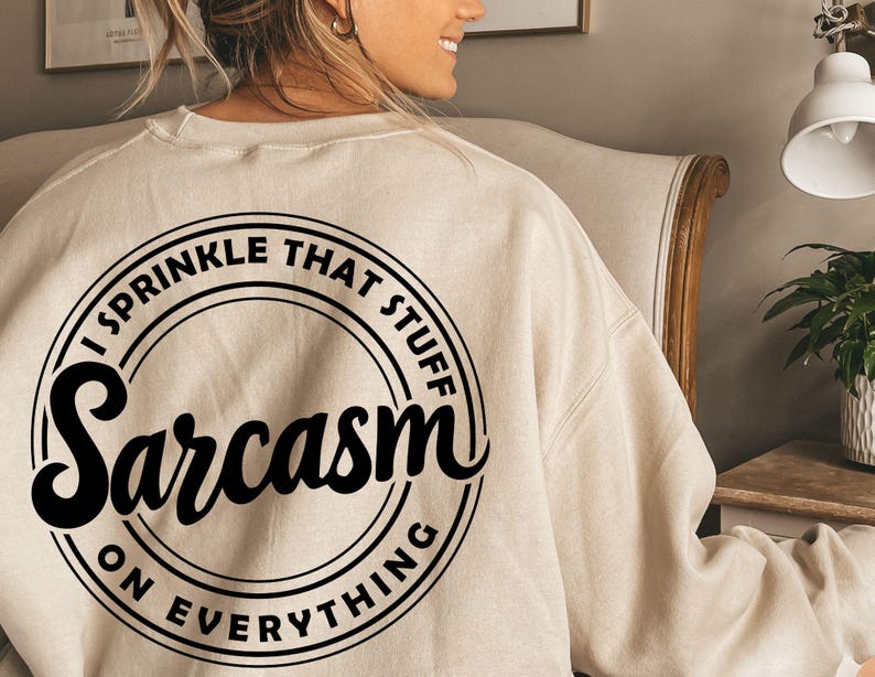 K&ouml;nnte beinhalten: Ein beiger Pullover mit einem schwarzen Kreis-Motiv, auf dem "I sprinkle that stuff Sarcasm on everything" steht.