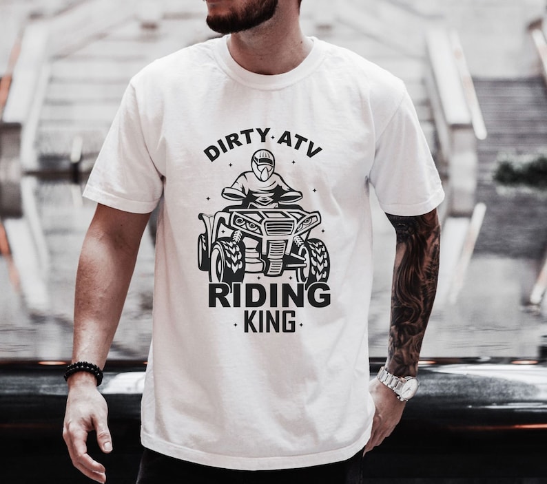 May include: White t-shirt with a black graphic of a person riding an ATV. The text "DIRTY ATV RIDING KING" is printed on the shirt.