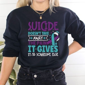 以下が含まれることがあります： 紫とターコイズブルーのリボンと「Suicide doesn't take away the pain it gives it to someone else」というテキストが書かれた黒いスウェットシャツ。