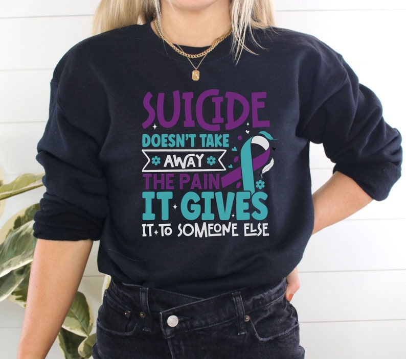以下が含まれることがあります： 紫とターコイズブルーのリボンと「Suicide doesn't take away the pain it gives it to someone else」というテキストが書かれた黒いスウェットシャツ。