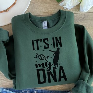 May include: A dark green sweatshirt with the text "It's in my DNA" in black lettering. A silhouette of a basketball player is shown next to the text. The text is surrounded by small stars.