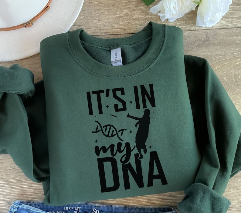 May include: A dark green sweatshirt with the text "It's in my DNA" in black lettering. A silhouette of a basketball player is shown next to the text. The text is surrounded by small stars.
