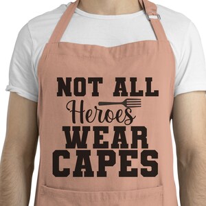 May include: A pink apron with adjustable straps and a large pocket. The apron has black text that reads "Not All Heroes Wear Capes" with a black spatula graphic.