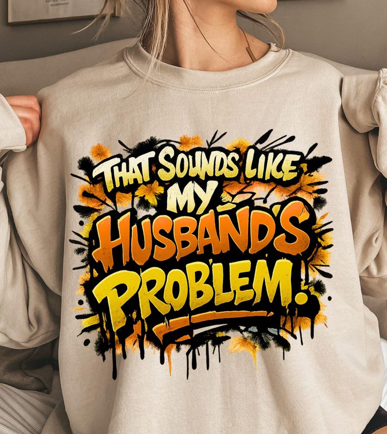 Pu&ograve; includere: Felpa beige con la scritta "THAT SOUNDS LIKE MY HUSBAND'S PROBLEM!" in un design in stile graffiti. Il testo &egrave; giallo e arancione, con dettagli neri e floreali.