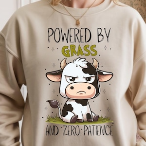 May include: Beige sweatshirt with a cartoon cow graphic. The cow is black and white with an angry expression, sitting on grass. Text above reads "POWERED BY GRASS" and below "AND ZERO PATIENCE".