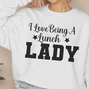 May include: White sweatshirt with black text that reads "I Love Being A *Lunch* Lady" with a star on either side of the word "Lunch".