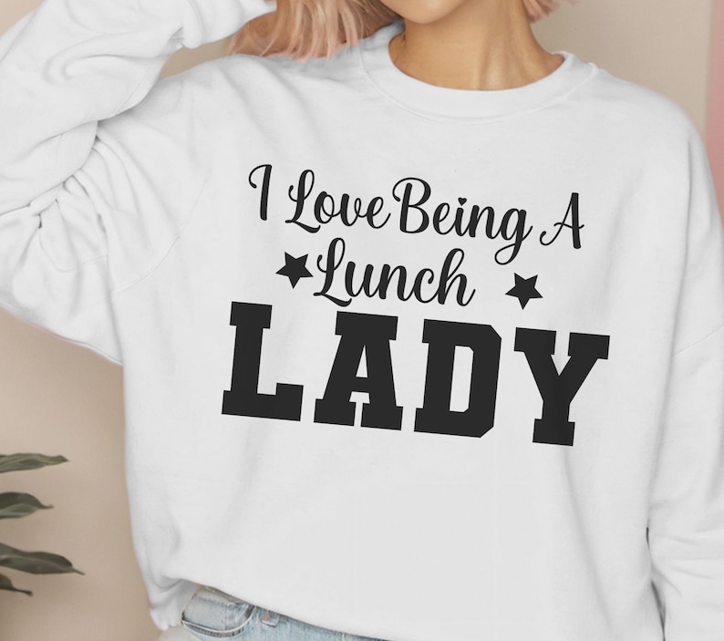 May include: White sweatshirt with black text that reads "I Love Being A *Lunch* Lady" with a star on either side of the word "Lunch".