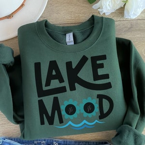May include: A dark green sweatshirt with the text "LAKE MOOD" in black lettering. The text is stylized with a wavy line underneath and two cartoon eyes with eyelashes.